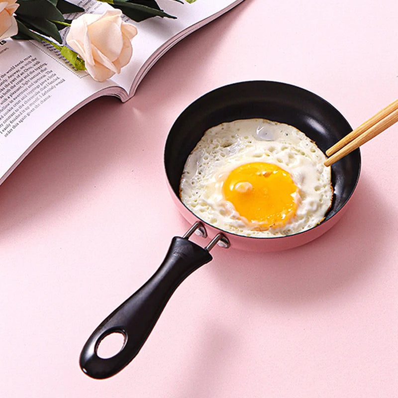 Small black frying pan with a fried egg and chopsticks on a pink surface with a magazine and flowers.