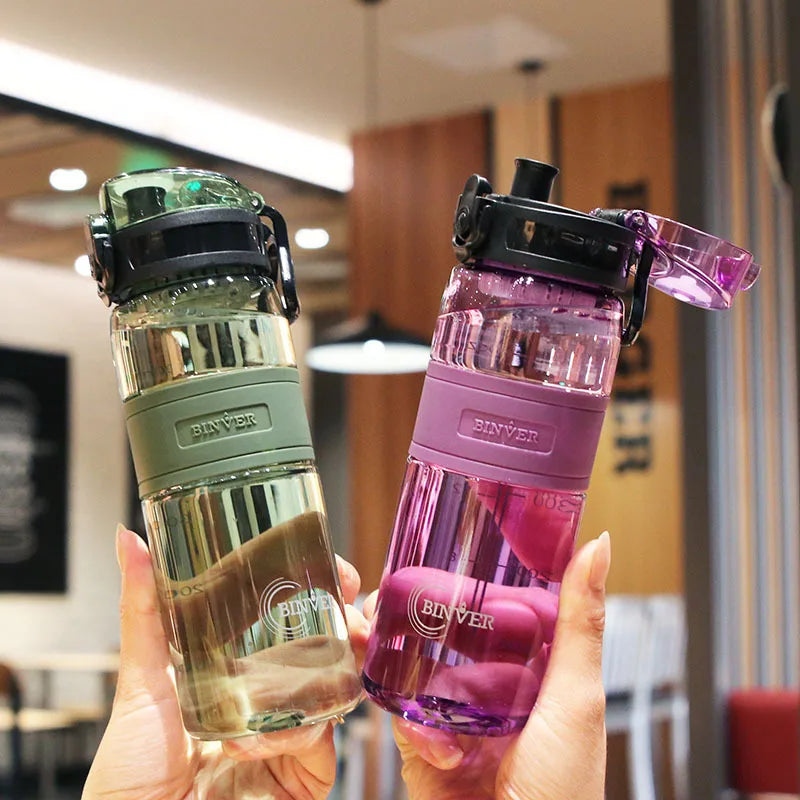 Two Biner water bottles, one green and one pink, held in a hand with a blurred indoor background.