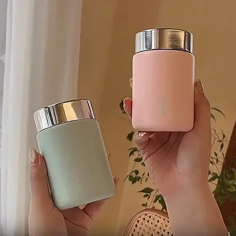 Two hands holding a light green and a pink container with silver lids against a neutral background.