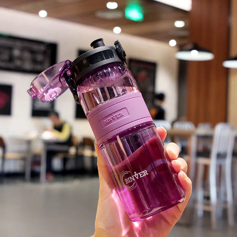 Pink water bottle with black cap held in a hand, blurred indoor background