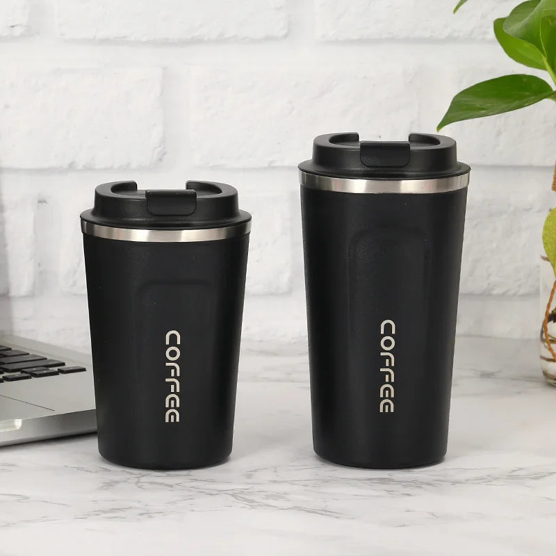 Two black coffee tumblers with silver lids on a marble surface next to a laptop.