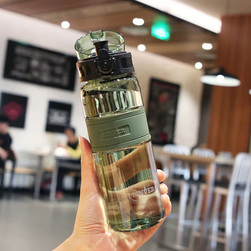 Hand holding a green water bottle with a blurred indoor setting in the background